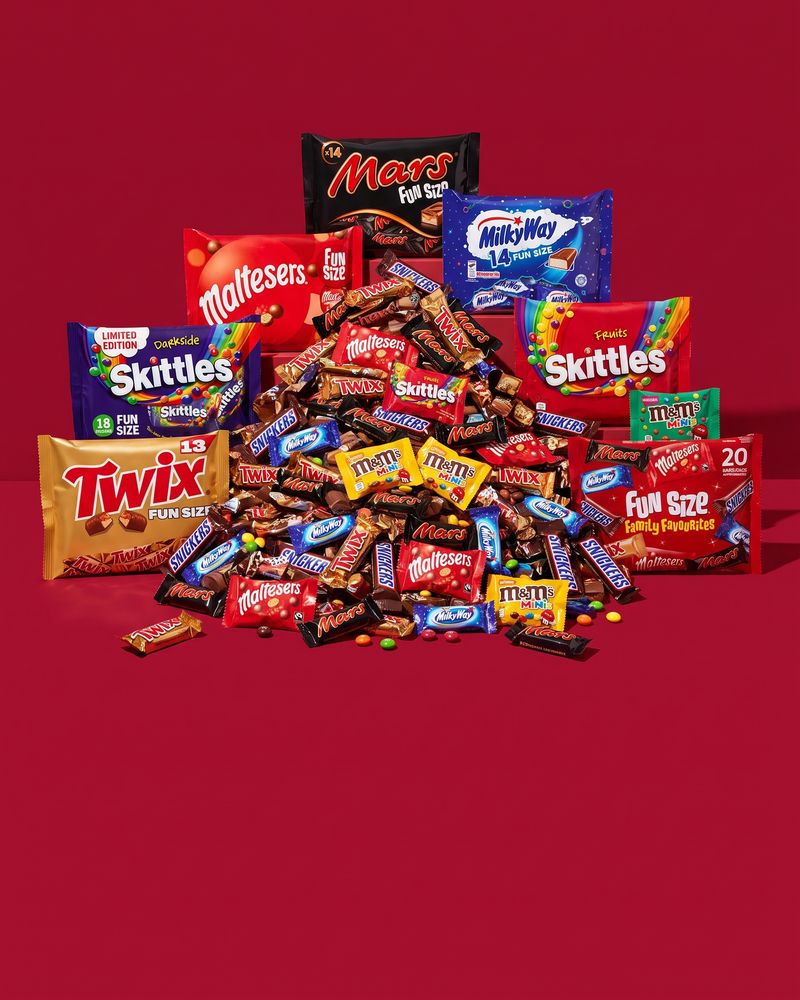 A heap of loose funsize chocolate bars on deep red, with Mars, Maltesers, Twix, Milky Way, Skittles and M&M's Minis sharing bags standing around it.