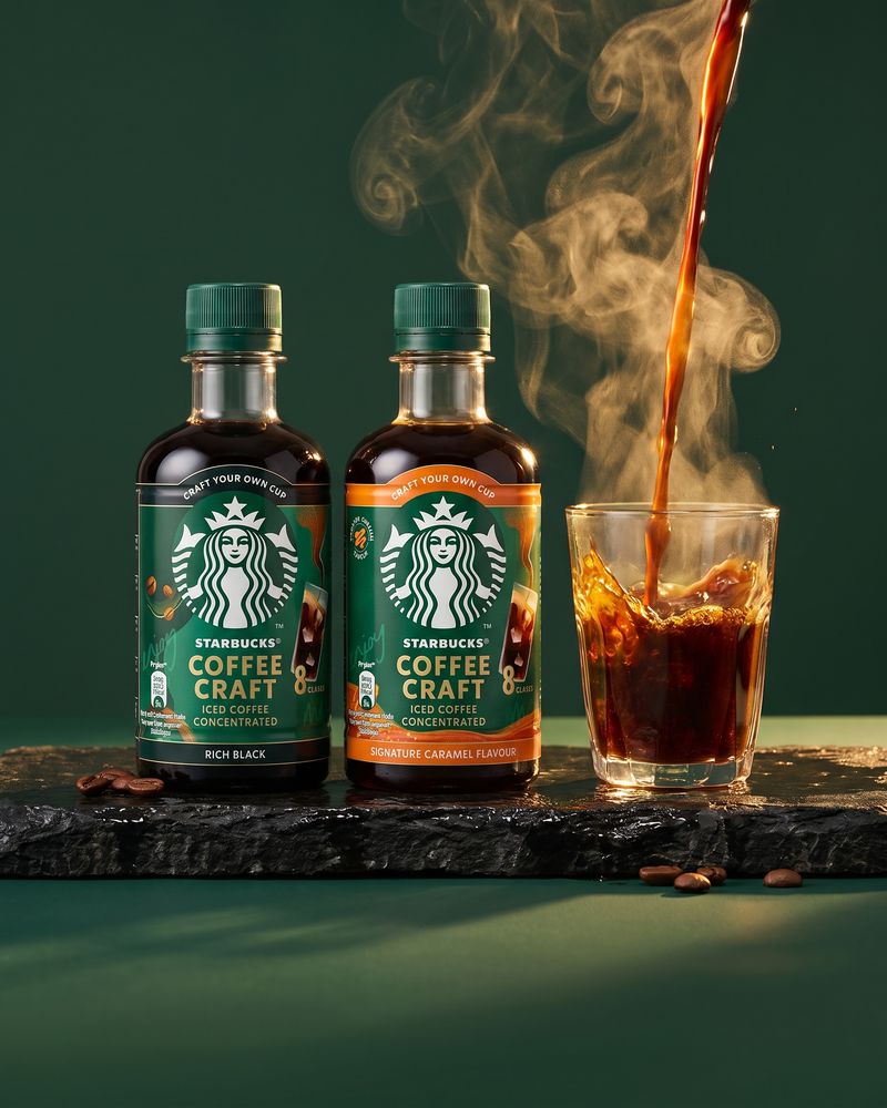 Two Starbucks Coffee Craft espresso concentrate bottles, Rich Black and Signature Caramel, on a wet slate ledge beside a glass taking a pour of coffee.